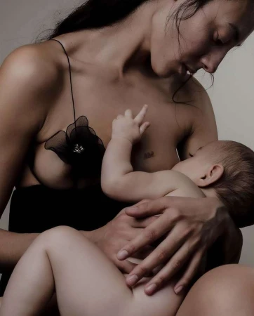 A mother nurses her infant in a close-up shot. The mother, wearing a black dress, looks down at her baby, the image is softly lit.