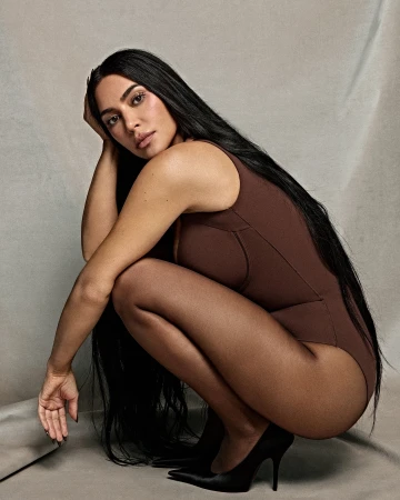 Brunette woman in a brown bodysuit and black heels posing in a photo studio, kneeling on a neutral gray backdrop with soft, diffused lighting.