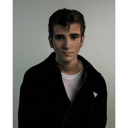 Portrait of young man with styled dark hair wearing black zip jacket and white t shirt against a grey background by Jet Swan