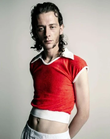 Man with dark curly hair wearing a red sleeveless collared top against a plain light background