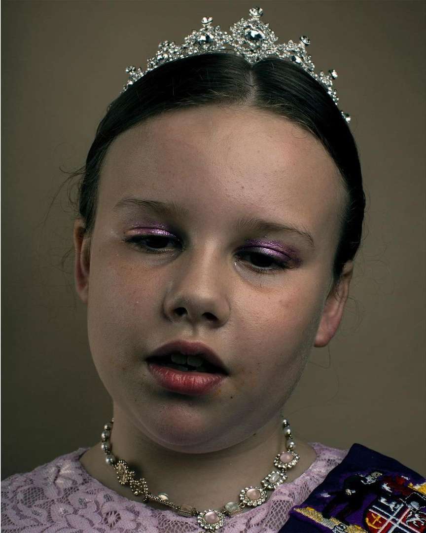 Portrait of a young girl with a tiara, necklace, and purple sash, wearing makeup and a lace top against a solid background.