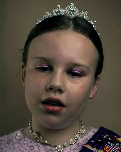 Portrait of a young girl with a tiara, necklace, and purple sash, wearing makeup and a lace top against a solid background.
