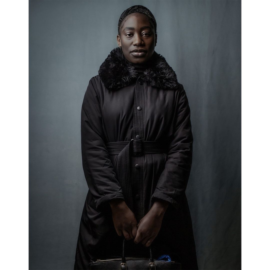 A Black woman stands indoors in a long black coat with fur trim, holding a black handbag, captured in a portrait-style photograph.