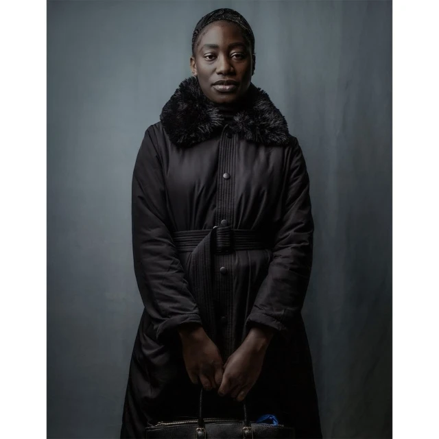 A Black woman stands indoors in a long black coat with fur trim, holding a black handbag, captured in a portrait-style photograph.