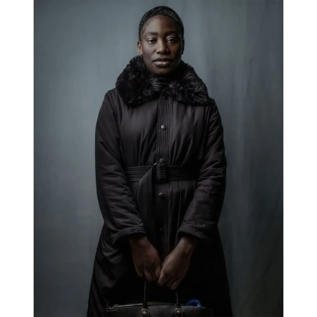 A Black woman stands indoors in a long black coat with fur trim, holding a black handbag, captured in a portrait-style photograph.