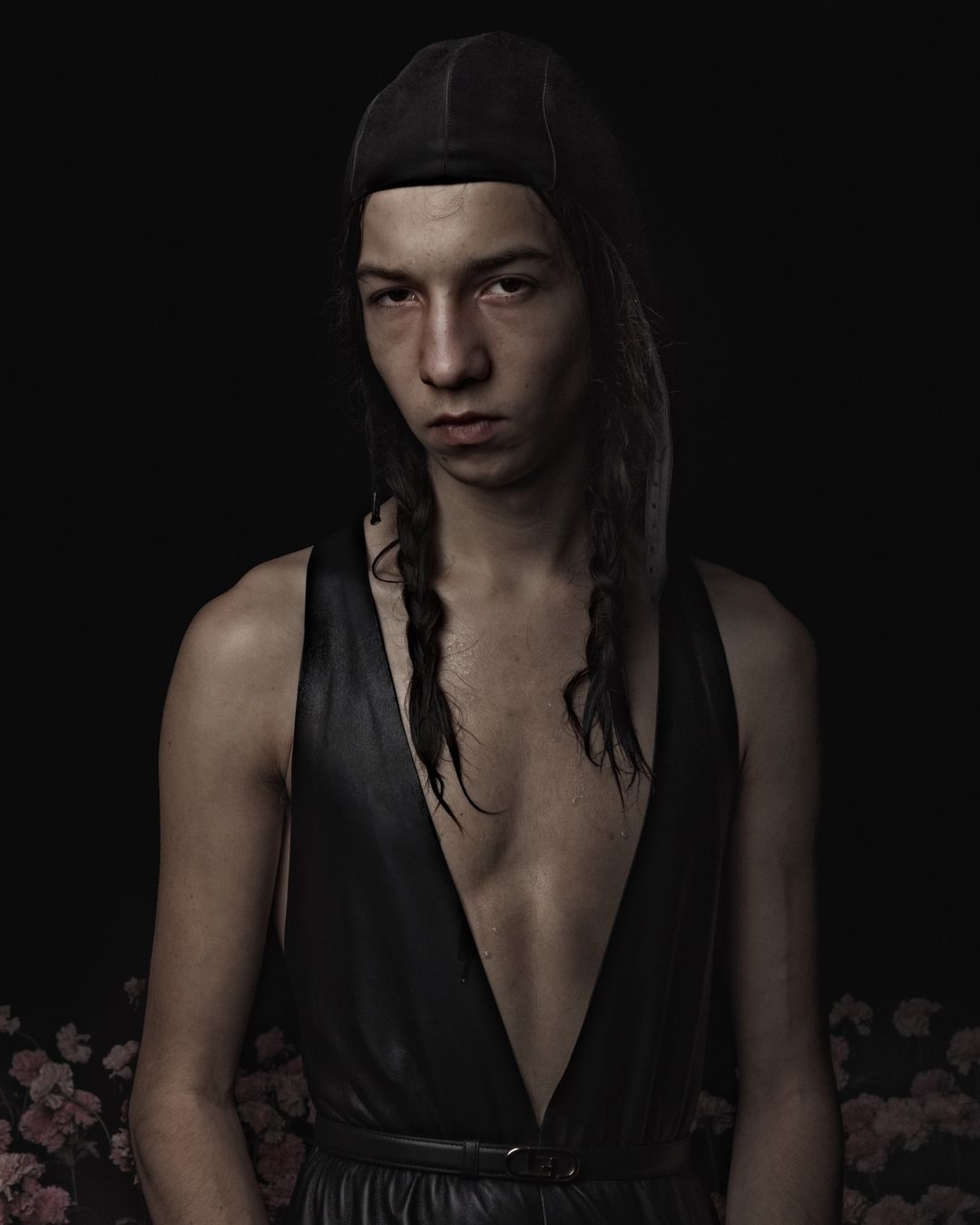 Portrait of a person with long braided hair wearing a sleeveless, low-cut black garment, black cap, against a dark background with pink flowers.