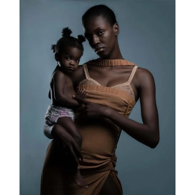 Studio shot of an African-American woman in a dress holding a baby in a diaper against a blue-grey background.