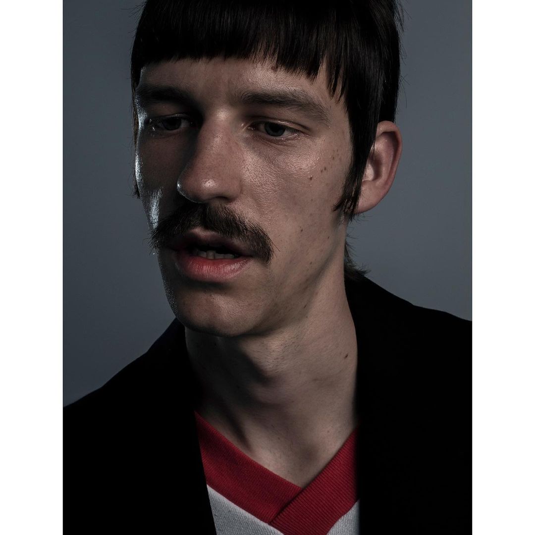 Close-up studio portrait of a fair-skinned man with a mustache and bangs, wearing a black jacket and red and white shirt.