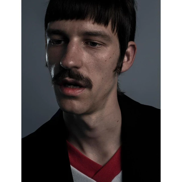 Close-up studio portrait of a fair-skinned man with a mustache and bangs, wearing a black jacket and red and white shirt.
