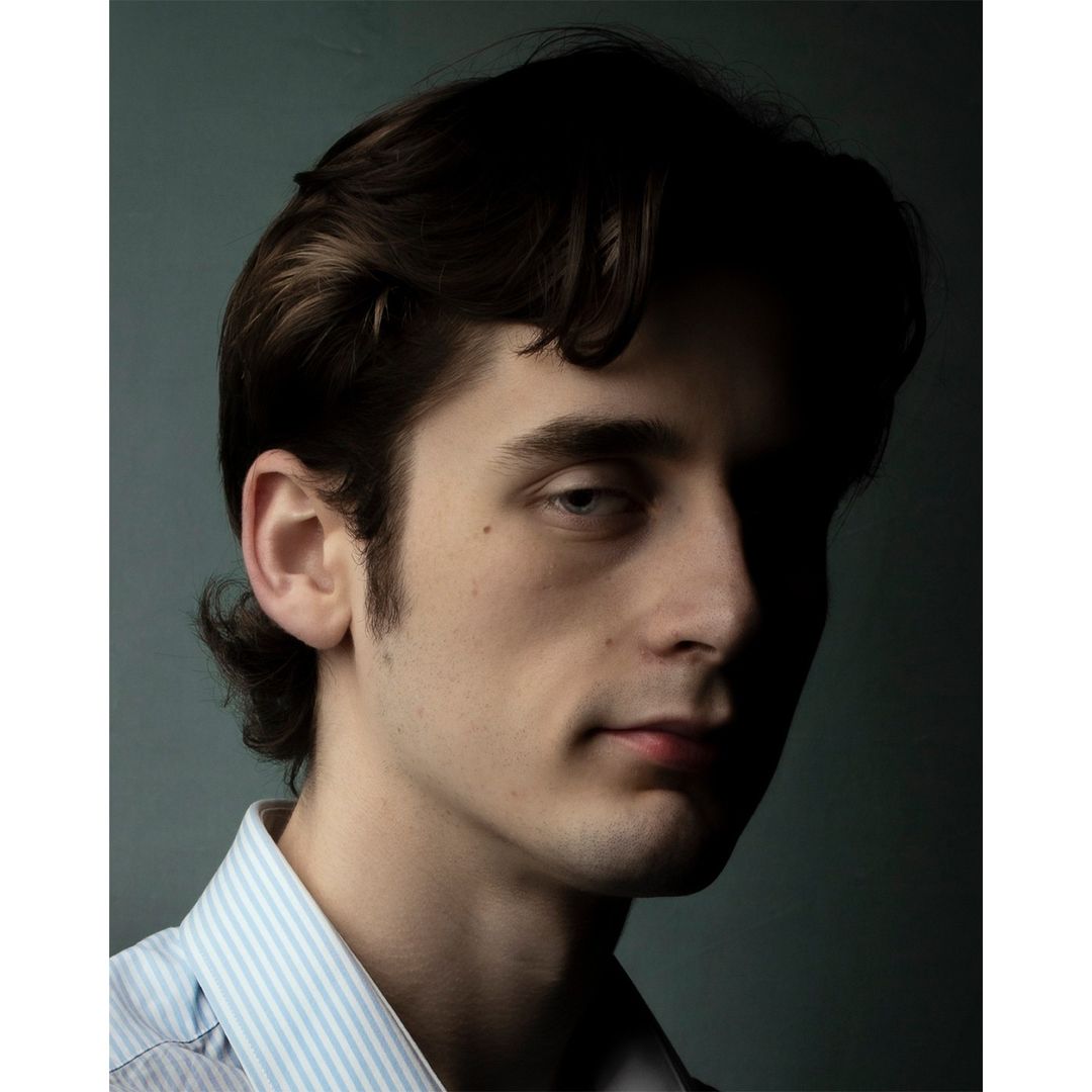 Dramatic portrait of a dark-haired young man in a blue-striped shirt, partially shadowed against a deep turquoise background.