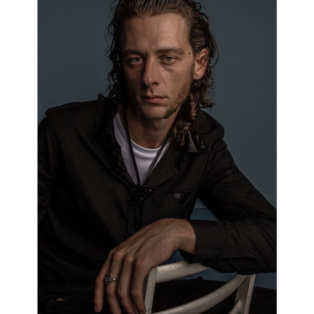 Portrait of a man with dark curly hair wearing a black shirt over a white t-shirt, seated on a white chair in London.