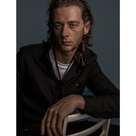Portrait of a man with dark curly hair wearing a black shirt over a white t-shirt, seated on a white chair in London.