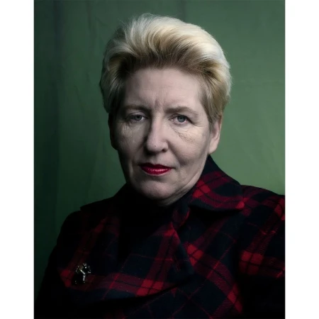 Portrait of an older woman with blonde hair wearing a red and black plaid jacket against a green backdrop.