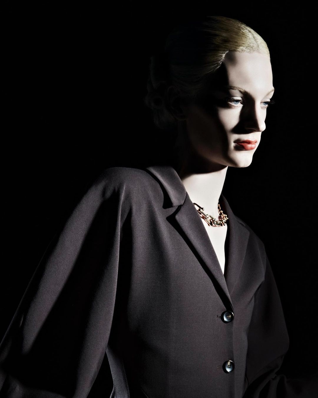 A mannequin in a black blazer and gold necklace is set against a dark background, illuminated with dramatic lighting.