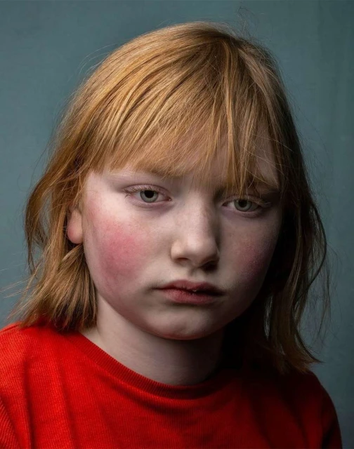 Portrait of a child with fair skin and red hair, wearing a red shirt, against a teal background in a close-up studio shot.
