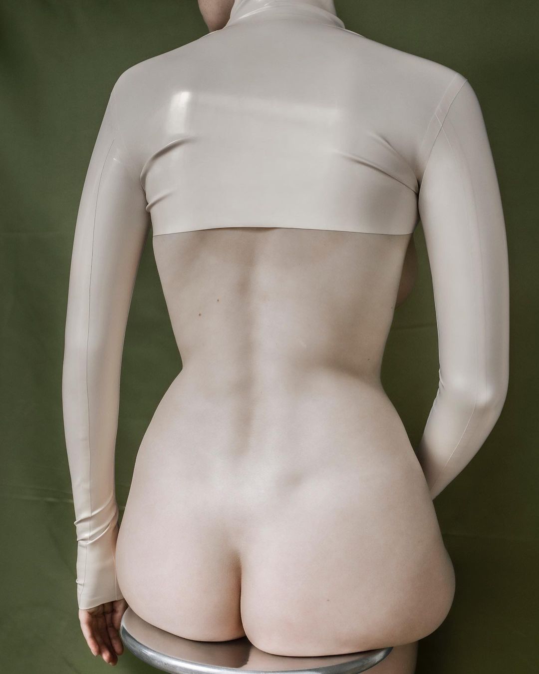 Back view of a slender adult in shiny beige latex top, sitting on a metal stool in front of an olive-green backdrop, medium shot.