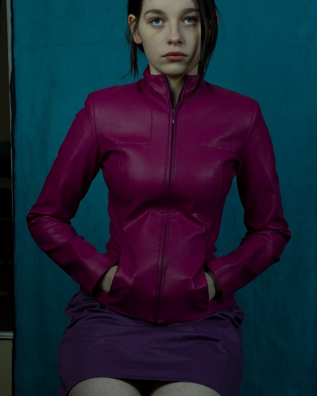 Young woman in magenta leather jacket poses against teal background. Fashion portrait with hands in pockets and dark hair.