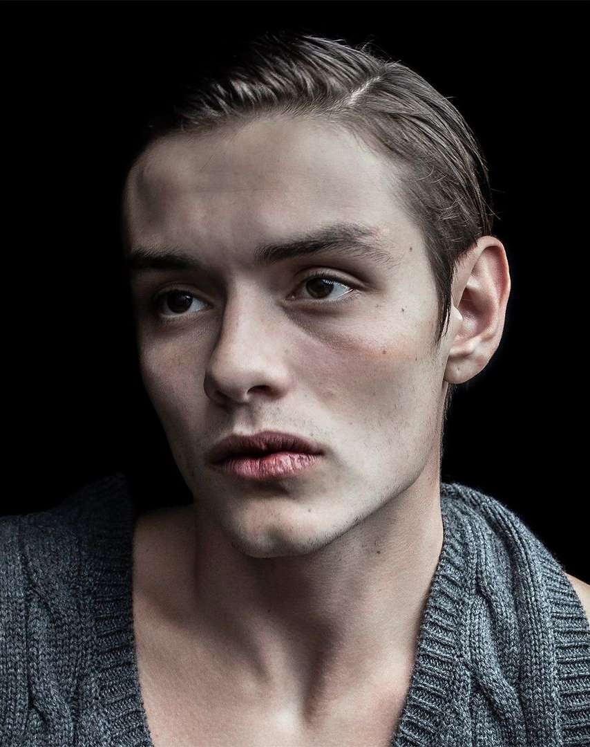Close-up portrait of a young man wearing a grey knitted tank top against a dark background by Jet Swan