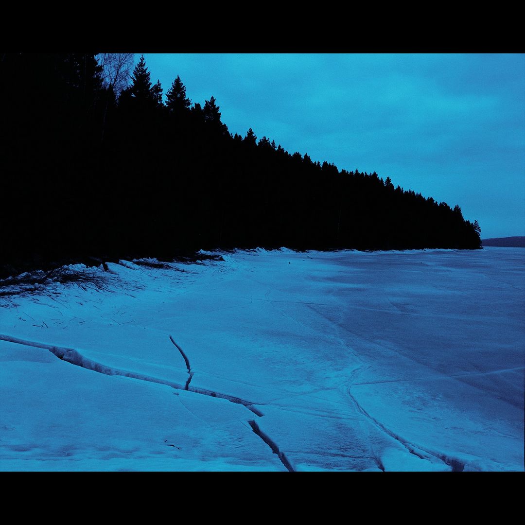 Landscape image featuring a frozen lake with cracked ice formations, silhouetted trees lining the shore, and a dusk sky, all under a blue color grade.