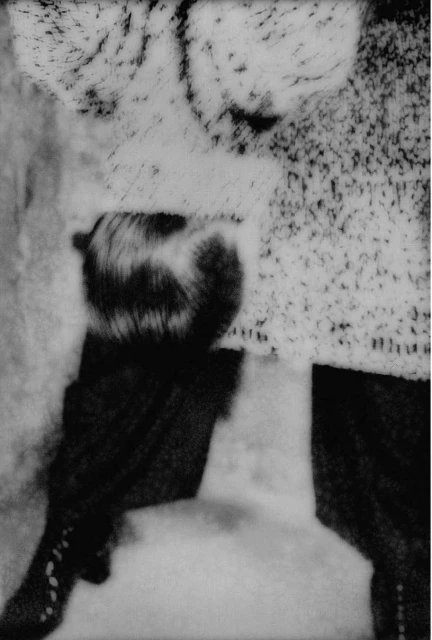 Black and white image of a woman with dark hair and a patterned shirt, set against a diffused background in a blurred indoor setting.