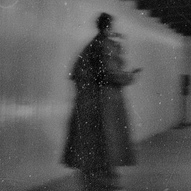 Monochrome image of a blurry, silhouetted person in a dark coat, possibly holding an object, set against a grainy background with stairs.