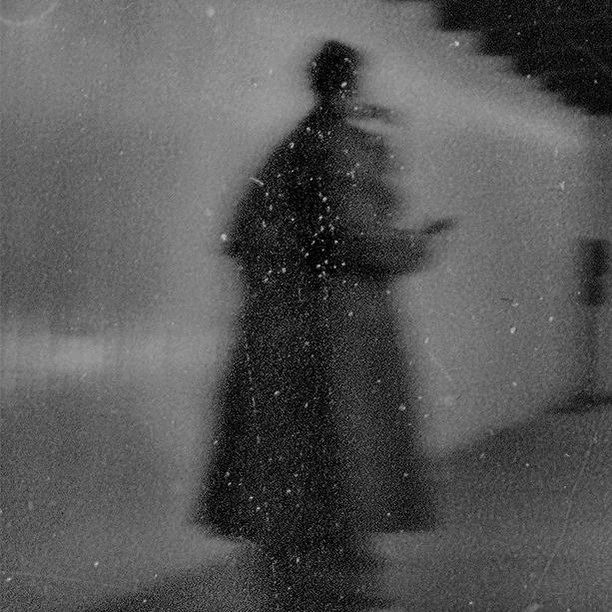 Monochrome image of a blurry, silhouetted person in a dark coat, possibly holding an object, set against a grainy background with stairs.