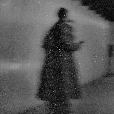 Monochrome image of a blurry, silhouetted person in a dark coat, possibly holding an object, set against a grainy background with stairs.