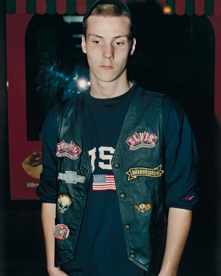 Young man posing in a leather vest with patches, wearing a black beanie and long-sleeved shirt, with an advertisement in the background.
