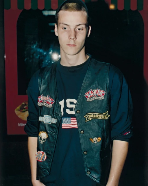 Young man posing in a leather vest with patches, wearing a black beanie and long-sleeved shirt, with an advertisement in the background.