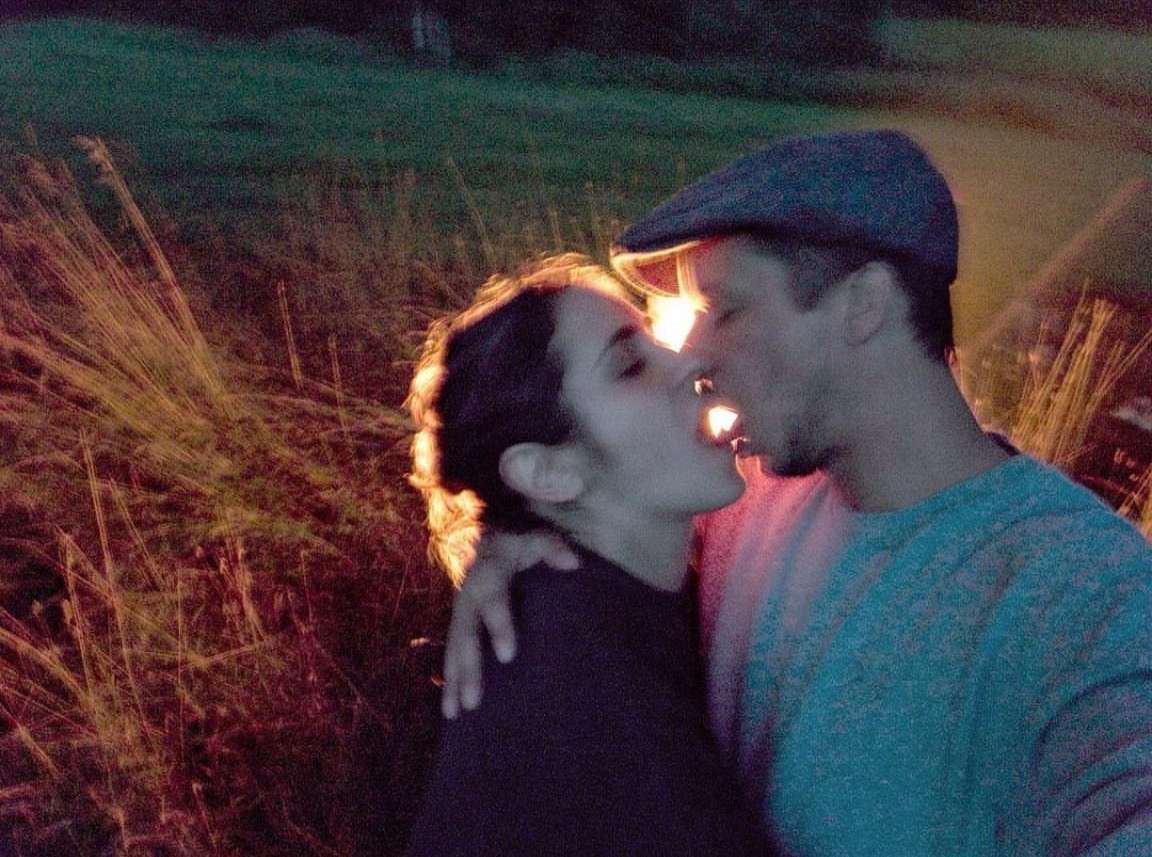 Close-up of a kissing couple in a field at dusk. The woman and man embrace in a romantic, outdoor environment with a grassy field.