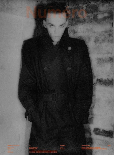 Fashion editorial image of a slender adult man in a long dark coat against a grey brick wall for Numero magazine cover, high contrast.