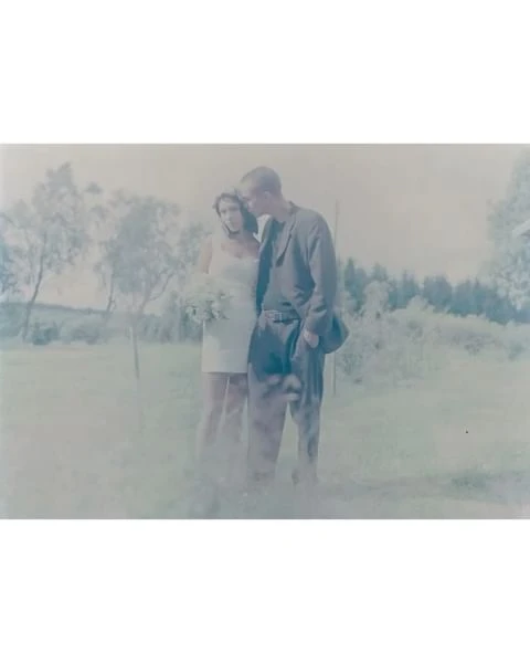 A fair-skinned couple poses in a field, the woman in a white dress holding flowers, and the man in a suit, on a cloudy day outdoors.