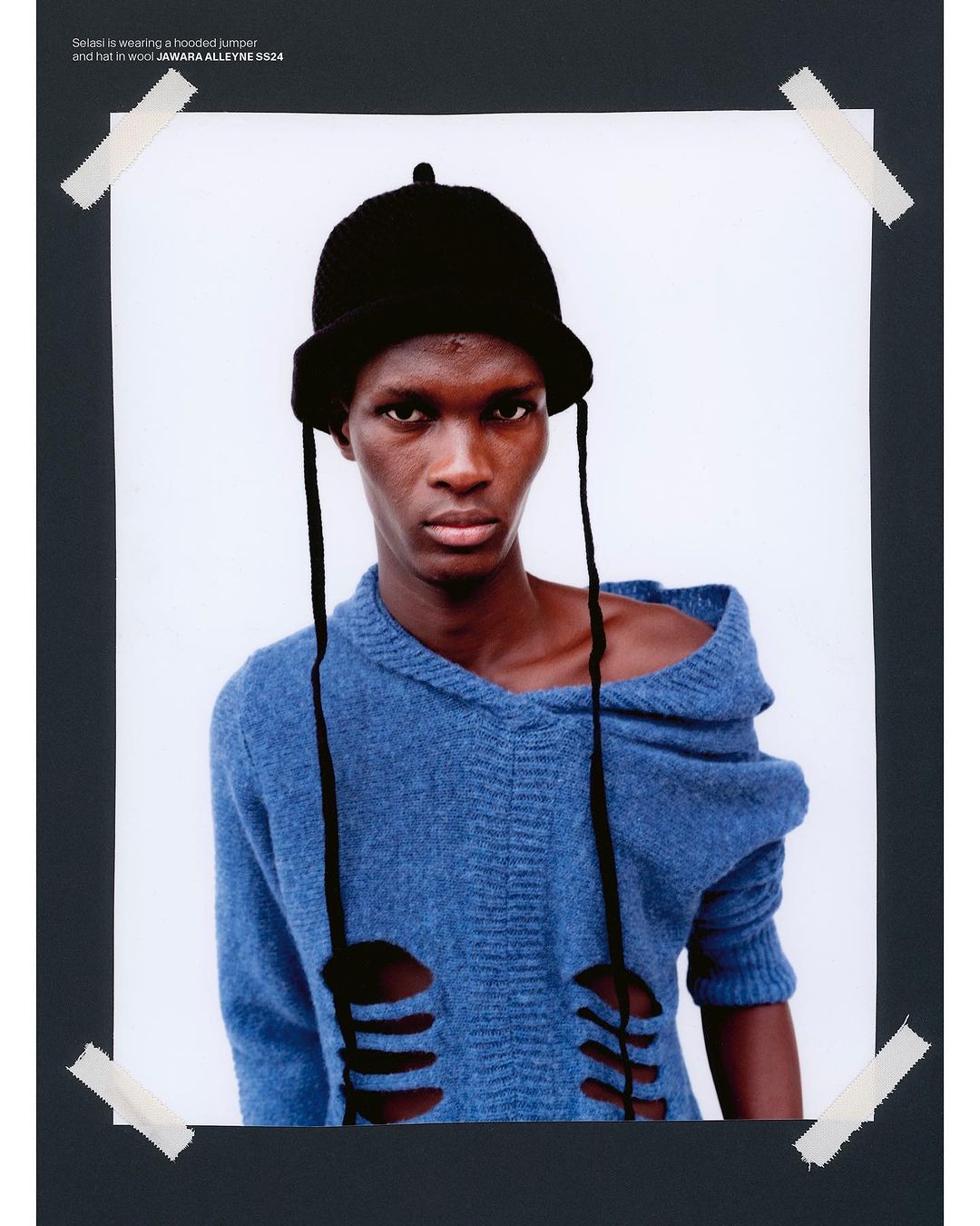 Close-up portrait of Black male model wearing a blue distressed hooded sweater and knit hat on a photo print taped to a dark background.