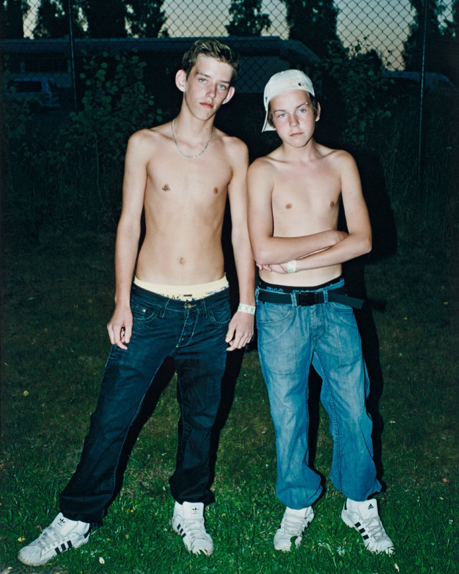 Two shirtless adolescent boys pose in jeans and sneakers in an outdoor setting. One wears a baseball cap; green foliage and a vehicle are visible in the background.