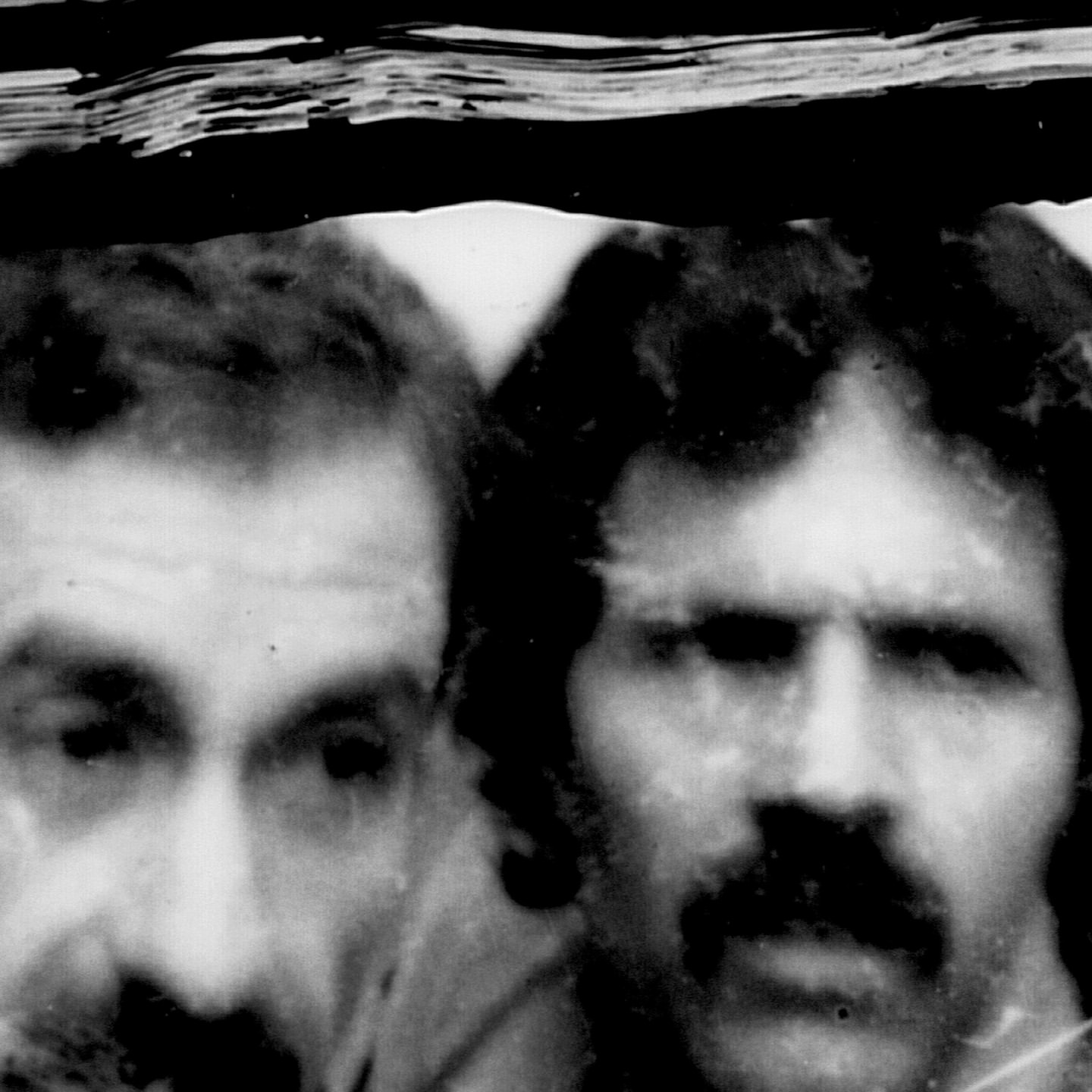 Black and white portrait of two adult males posing indoors. The image exhibits high contrast and a vintage aesthetic. One man has a mustache.