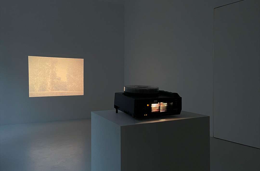 Slide projector on a pedestal in a darkened room, projecting an image of a landscape onto the wall, creating an art installation.
