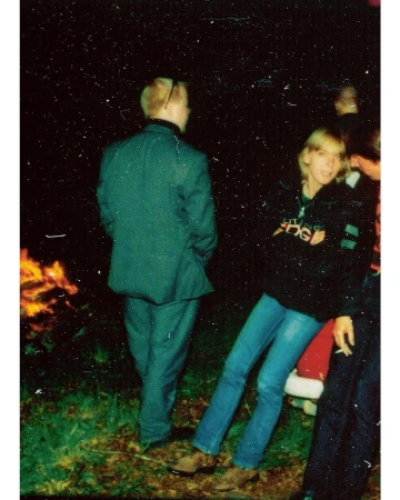 Group of people gathered around a fire at night, one man in a suit, another in jeans and a hoodie, at an outdoor event.