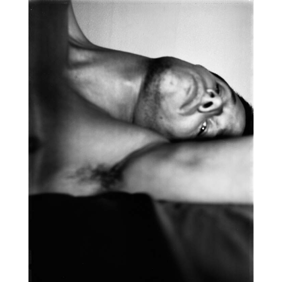 Black and white portrait of a man lying down, face towards the camera, with visible armpit hair against a neutral background.
