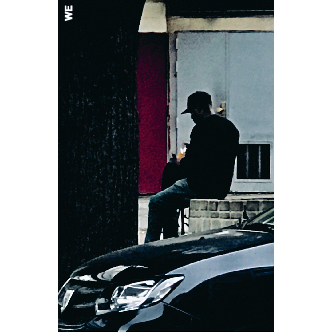 Adult male in baseball cap sits outdoors near tree and parked car; casual clothing, moody lighting, gray building backdrop.
