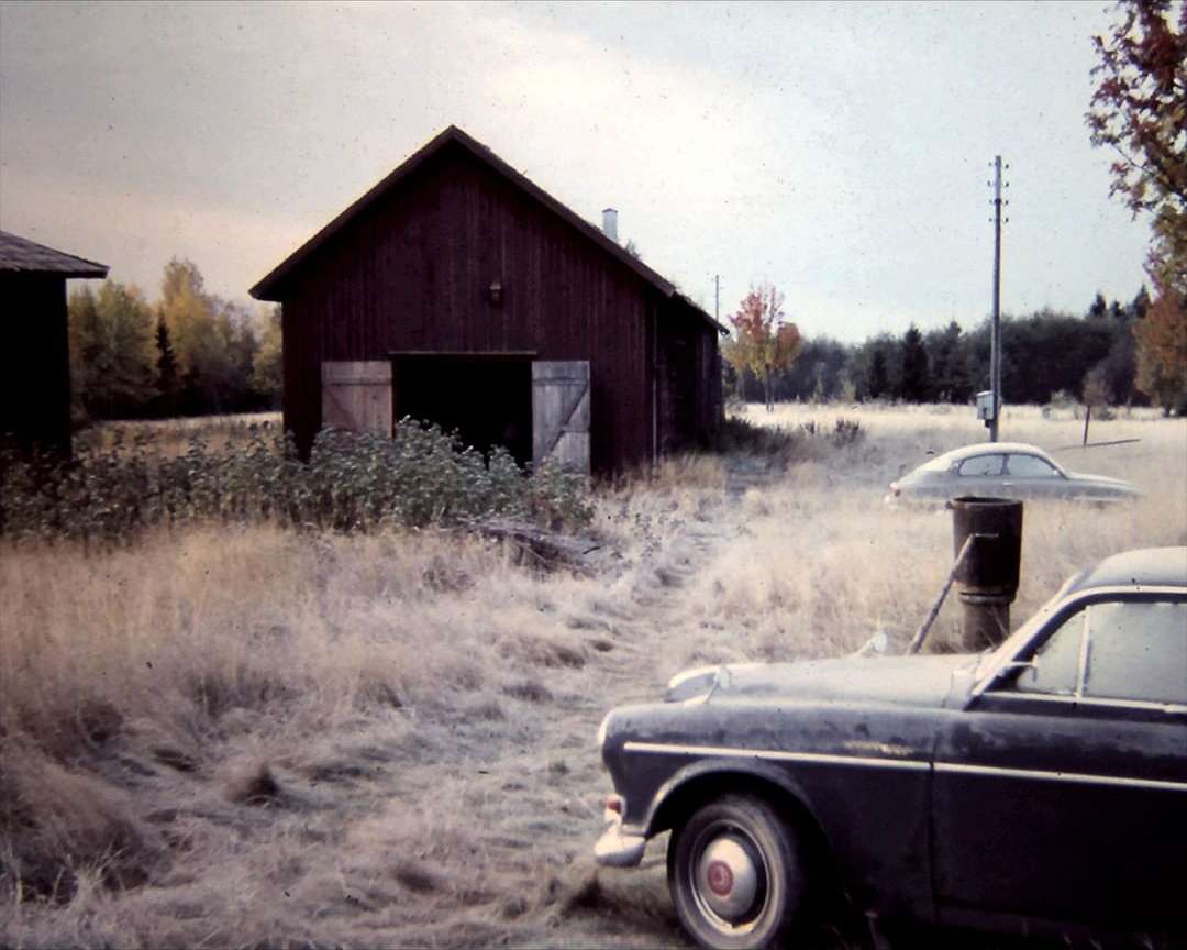 Vintage cars parked near a red barn in a rural landscape with tall grass and trees under an overcast sky, creating a nostalgic scene.