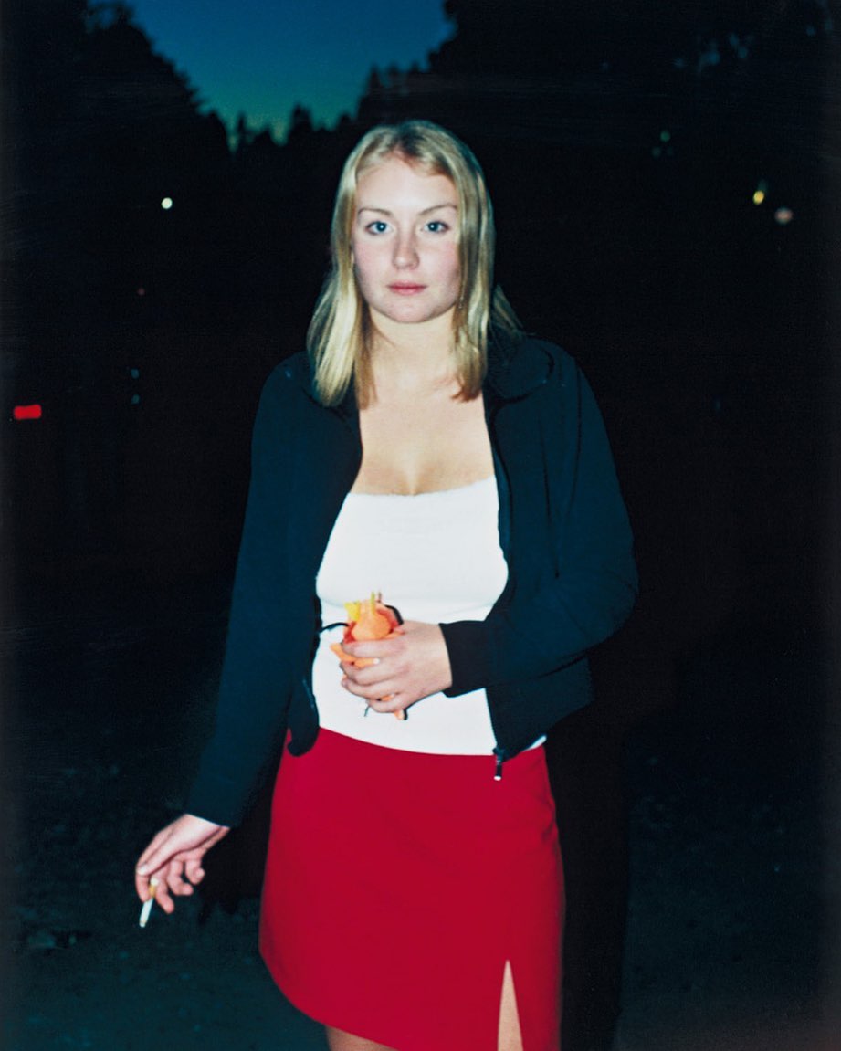 Portrait of a blonde woman in a black jacket and red skirt, holding an orange, standing outdoors at dusk, with a cigarette in her hand.