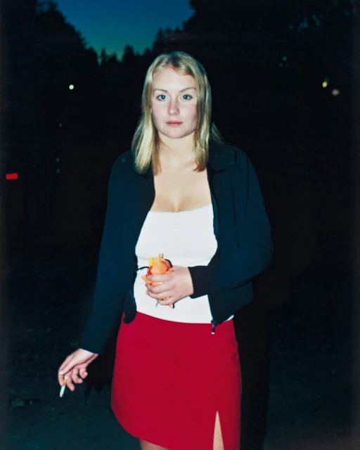 Portrait of a blonde woman in a black jacket and red skirt, holding an orange, standing outdoors at dusk, with a cigarette in her hand.