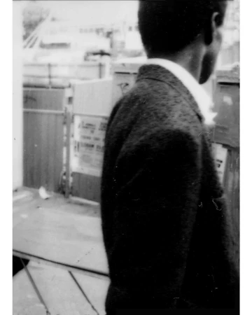 Monochrome portrait of an African American adult male in profile, wearing a jacket and shirt, set against an outdoor urban backdrop.