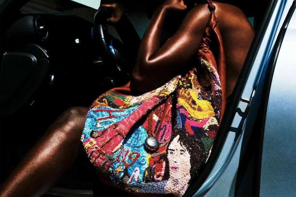 A dark-skinned woman is seated inside a car, a colorful, embellished purse hanging from her shoulder. Bright lighting accentuates the scene.