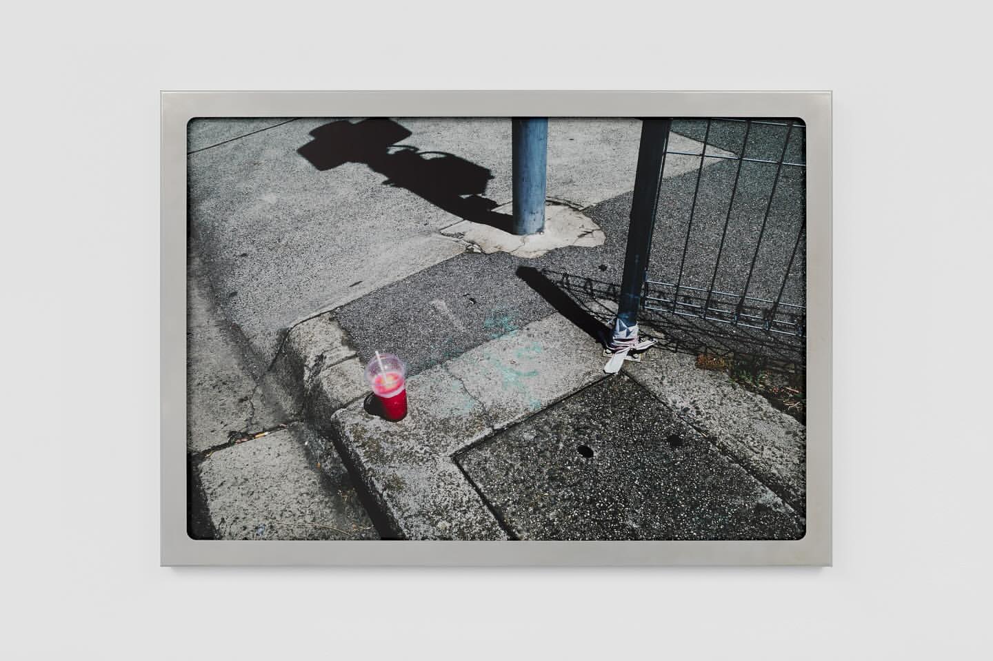 Framed photo of a red drink on a city sidewalk, with shadows of a street sign and light pole creating an urban still life.
