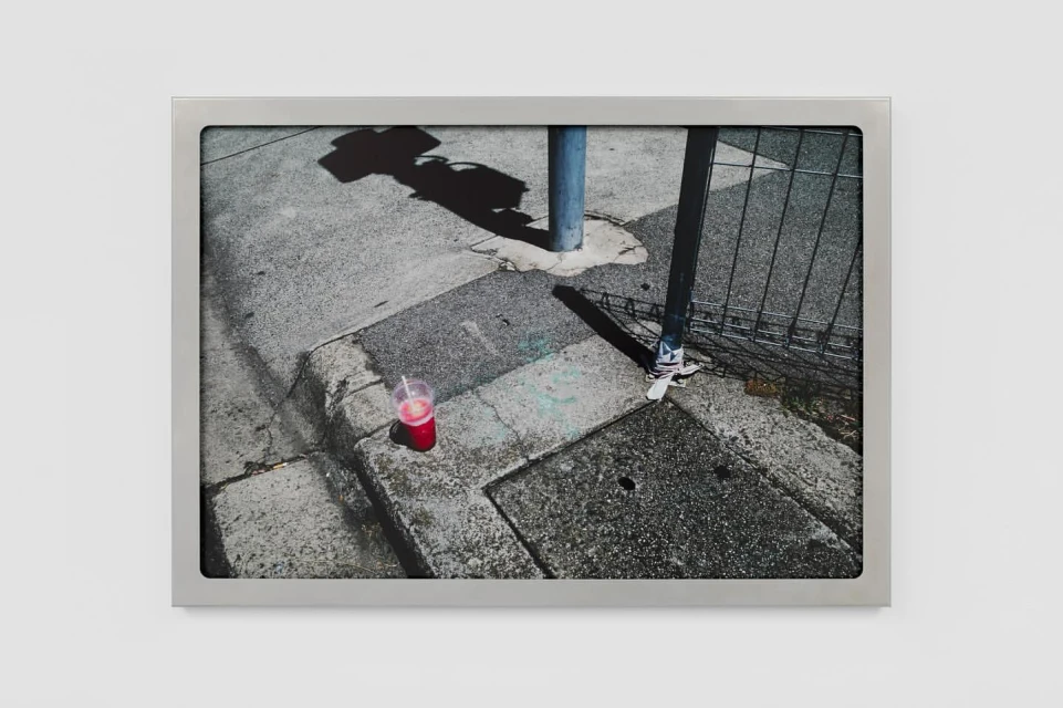 Framed photo of a red drink on a city sidewalk, with shadows of a street sign and light pole creating an urban still life.