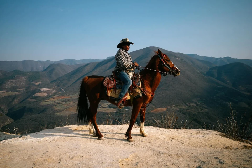A man riding a horse on a white ridge, with the background displaying a mountain range and clear sky in an outdoor setting.