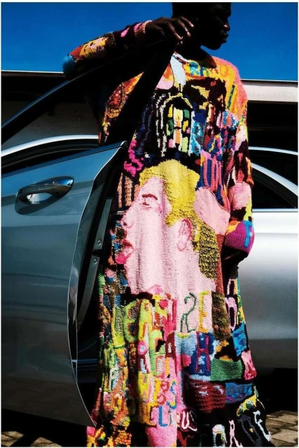 Dark-skinned adult wearing a colorful, patterned knit dress leaning against an open silver car under a bright blue sky outdoors.