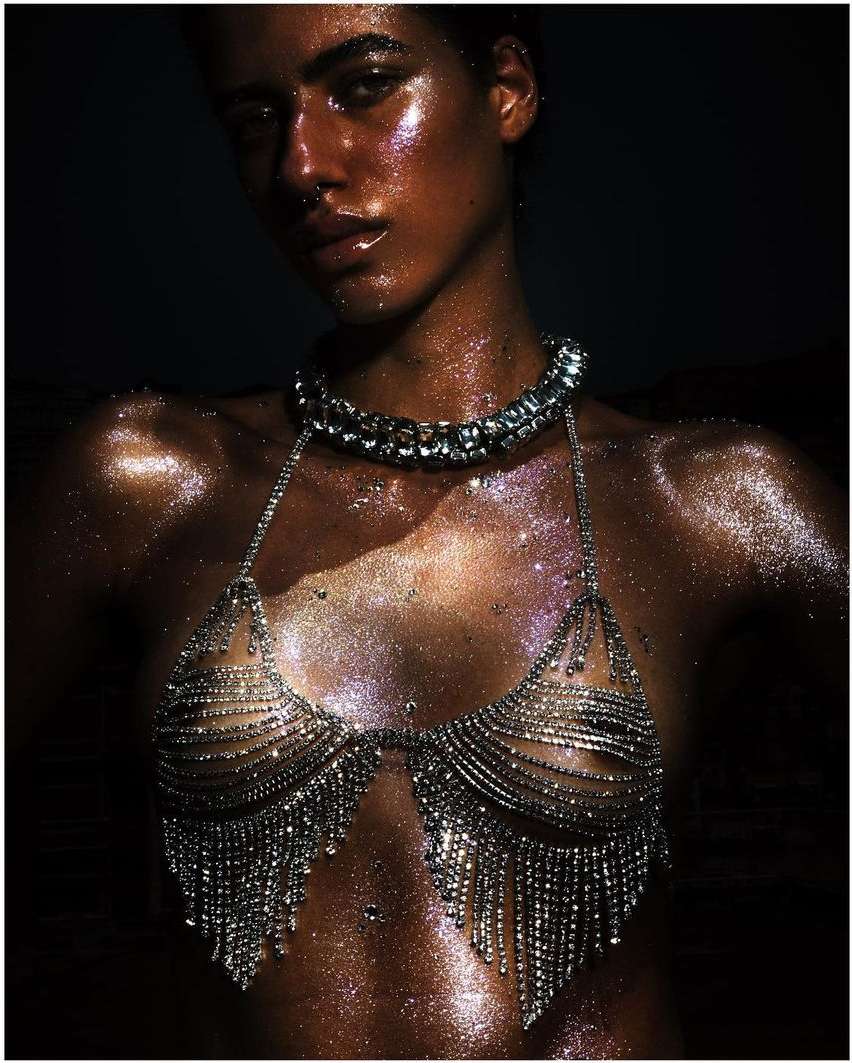 Close-up of a woman with shimmering bronze skin, wearing a jeweled bikini top and necklace, with a dark background in an indoor setting.