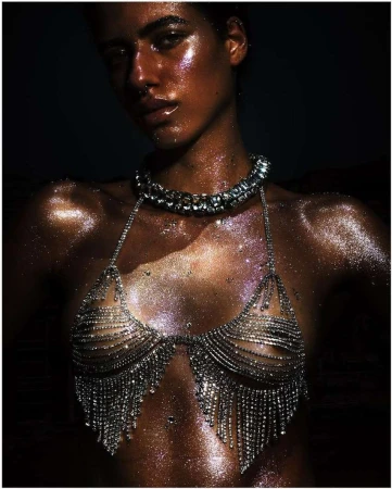 Close-up of a woman with shimmering bronze skin, wearing a jeweled bikini top and necklace, with a dark background in an indoor setting.